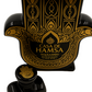 The Recycled Black and Gold Tequila Candle is crafted in the shape of a Hamsa hand, featuring intricate patterns, two candle-like flames, and Coconut Apricot wax, with the label "Casa de Hamsa Tequilanejo Hecho en México.