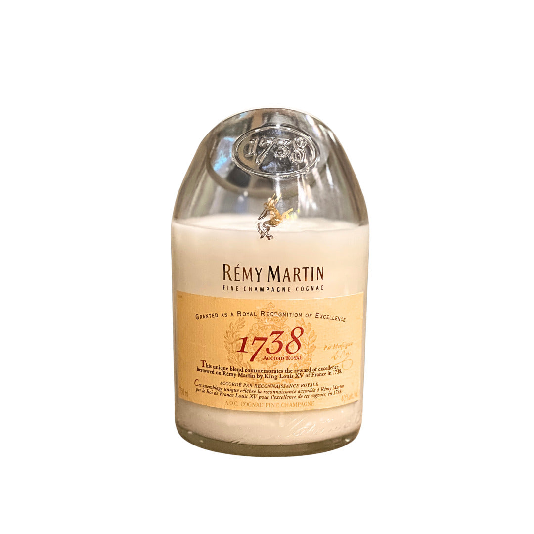 The Recycled Cognac Candle is a white candle in a glass container made from recycled cognac bottles, featuring a Rémy Martin 1738 label and topped with a metal medallion.