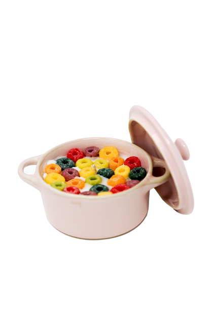 The Pink Mini Cereal Crockpot Candle, a small pink ceramic bowl with a partially open lid, is filled with colorful Froot Loops candle pieces and milk and sits on a white background.