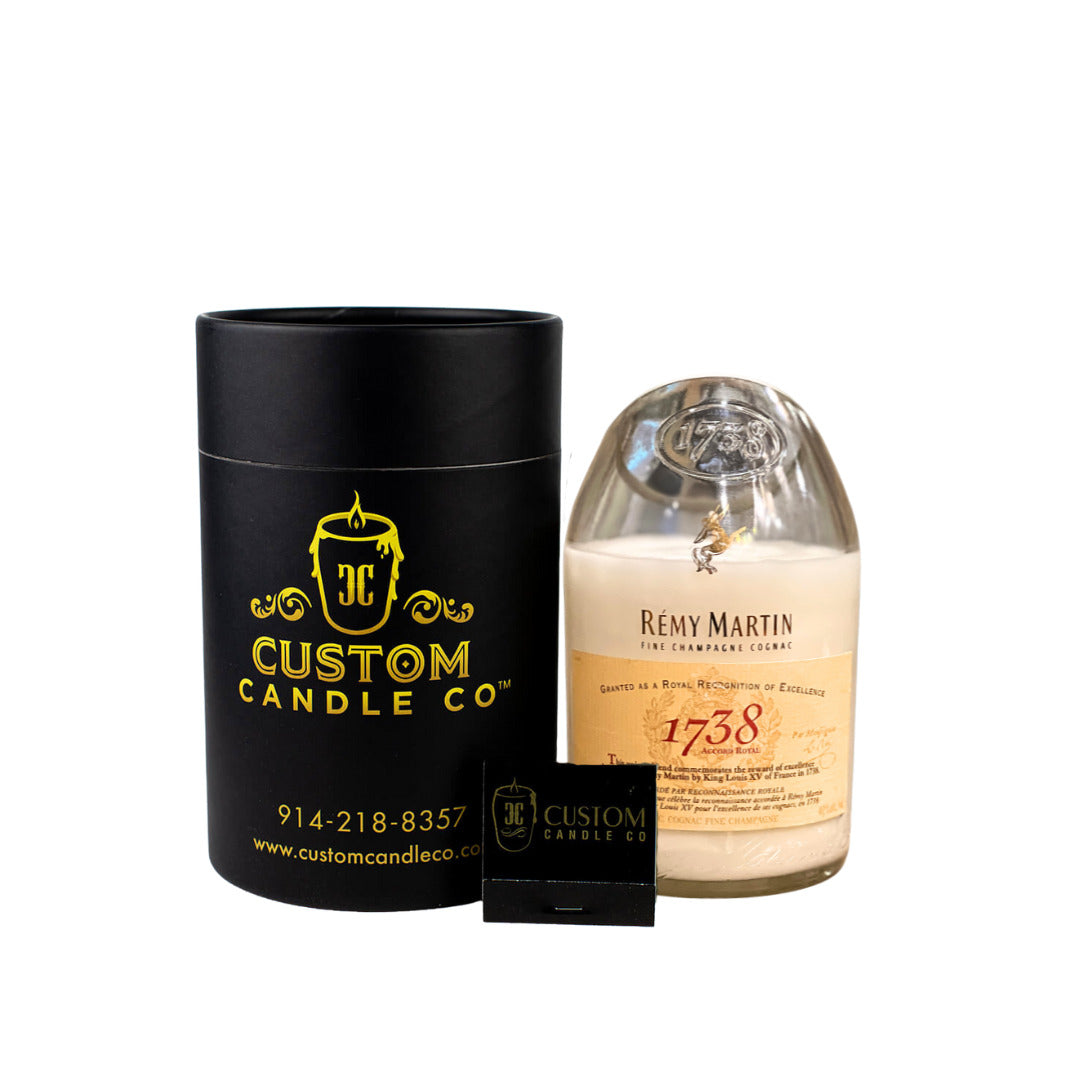 A Recycled Cognac Candle in a Rémy Martin 1738 bottle sits beside a black Custom Candle Co. container, with a business card displayed in front.