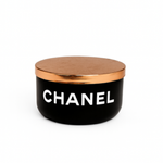 The Designer Noir Candle features a black round container with a metallic gold lid and "CHANEL" in white on the front, exuding elegance and sophistication.
