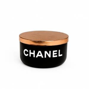 The Designer Noir Candle features a black round container with a metallic gold lid and "CHANEL" in white on the front, exuding elegance and sophistication.