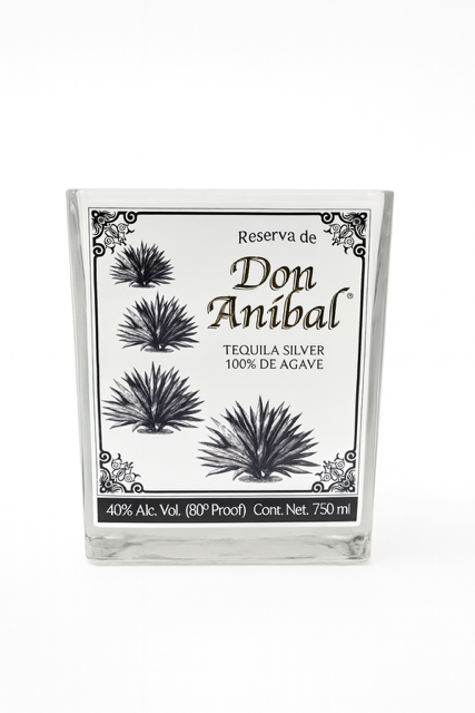 The Recycled Silver Tequila Candle features a clear glass bottle with black agave plant illustrations and a decorative border on the label, inspired by Don Anibal Silver Tequila (100% agave, 40% ABV, 750 ml).