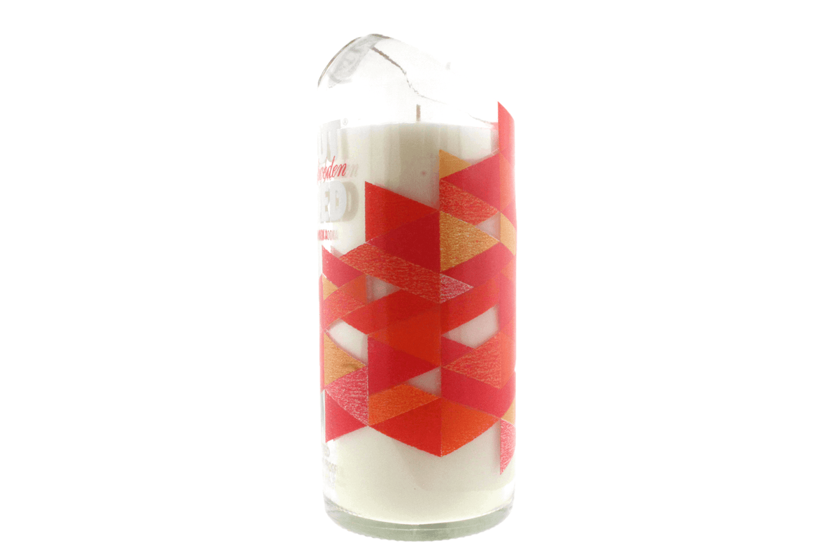 The Recycled Ruby Vodka Candle is a hand-poured natural soy candle in a recycled vodka glass with a red and orange geometric triangle design. The glass has a chipped top.