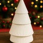 The White Ceramic Christmas Tree Candle, hand-poured from all natural wax, features three ribbed tiers and sits on a wooden surface glowing warmly amid blurred holiday decorations and festive lights in the background.
