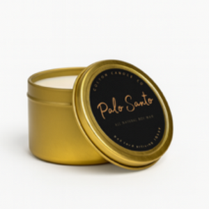 A gold-toned tin from the Palo Santo Box Set, with a partially open lid, displays a white candle inside.