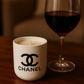 The White Ceramic Designer Candle sits beside a glass of red wine on a wooden surface, with a blurred background.