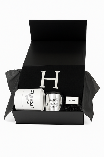 The Paris Designer Gift Set includes a black gift box with a black item featuring a white "H," a white Hermès candle, a silver Hermès cup, and a cotton throw—an elegant choice for those who value custom-designed gift baskets.