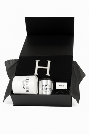 The Paris Designer Gift Set includes a black gift box with a black item featuring a white "H," a white Hermès candle, a silver Hermès cup, and a cotton throw—an elegant choice for those who value custom-designed gift baskets.