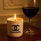 A White Ceramic Designer Candle glows beside a glass of red wine on a wooden table, set against an elegant, softly lit background.