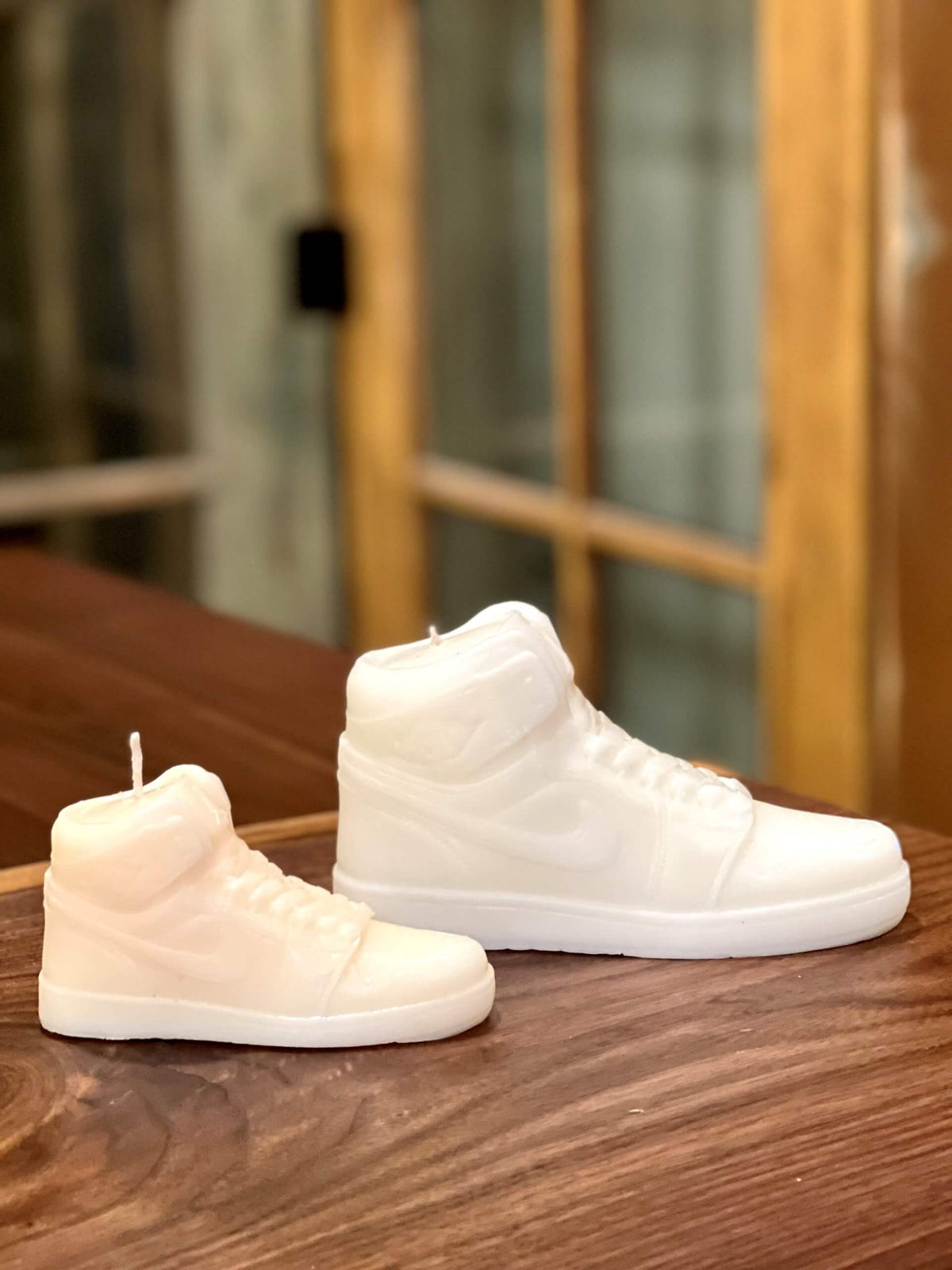 A small AJ-1 White Hightop Sneaker Candle, hand poured with all natural wax, rests on a wooden surface before blurred windows; this product is currently on backorder.