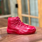 The AJ-11 Red Hightop Sneaker Candle – Large (On Backorder), hand-poured with all-natural wax, sits on a wooden surface with a blurred background of windows and wood paneling.