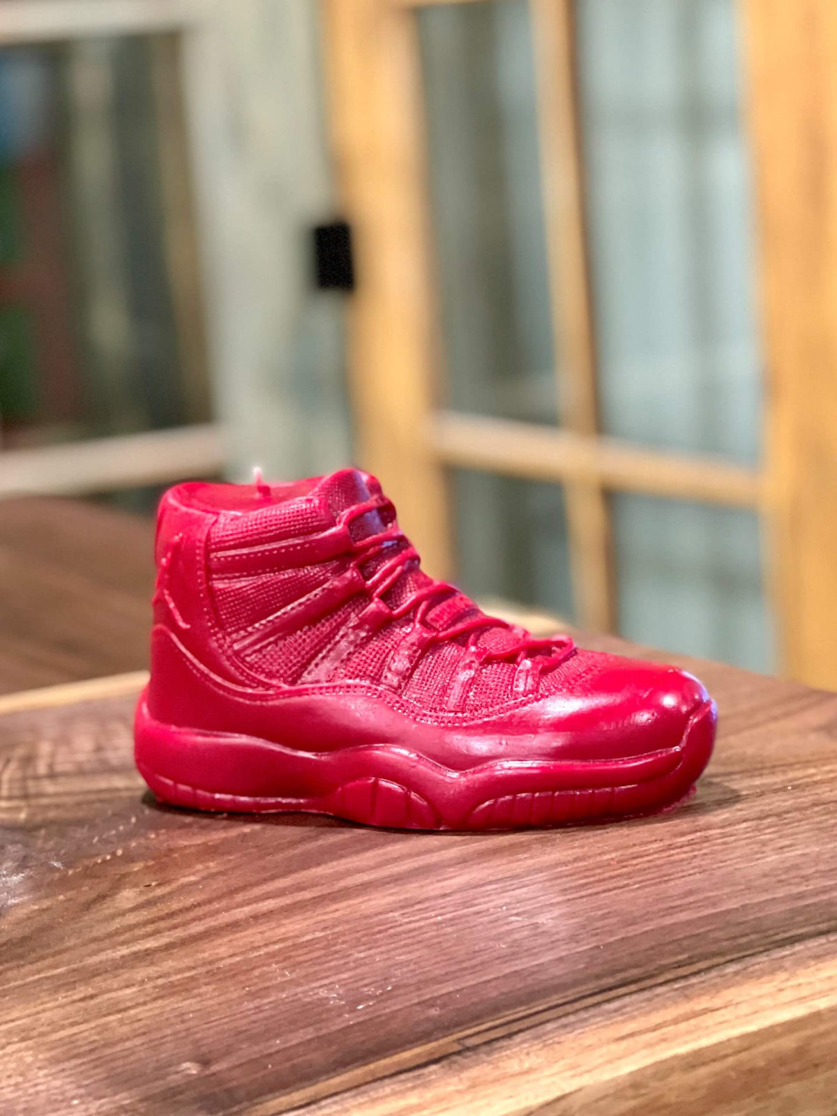 The AJ-11 Red Hightop Sneaker Candle – Large (On Backorder), hand-poured with all-natural wax, sits on a wooden surface with a blurred background of windows and wood paneling.