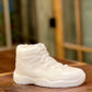 The AJ-11 White Hightop Sneaker Candle – Large (On Backorder), a hand-poured candle shaped like a white high-top sneaker, is displayed on a wooden surface with a blurred window in the background.