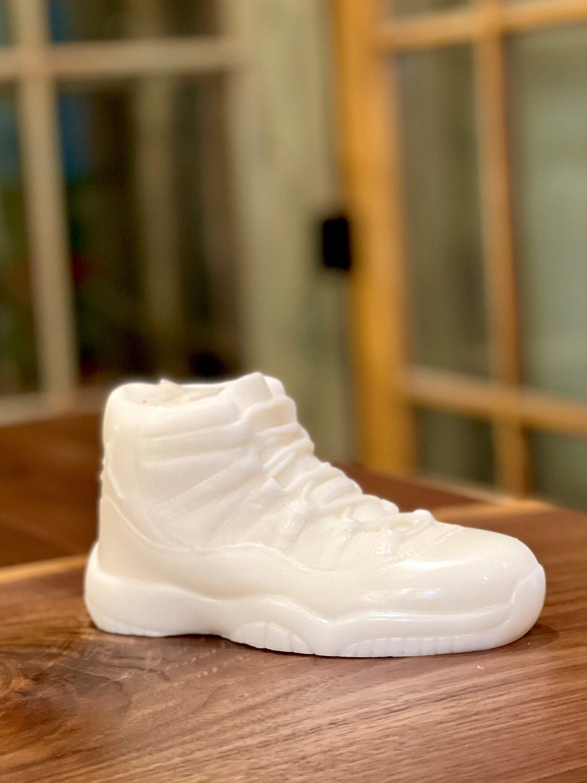 The AJ-11 White Hightop Sneaker Candle – Large (On Backorder), a hand-poured candle shaped like a white high-top sneaker, is displayed on a wooden surface with a blurred window in the background.