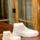 A large AJ-11 White Hightop Sneaker Candle sits on a wooden surface by glass-paneled doors. Hand poured to capture every detail, this candle adds a stylish accent to any space.