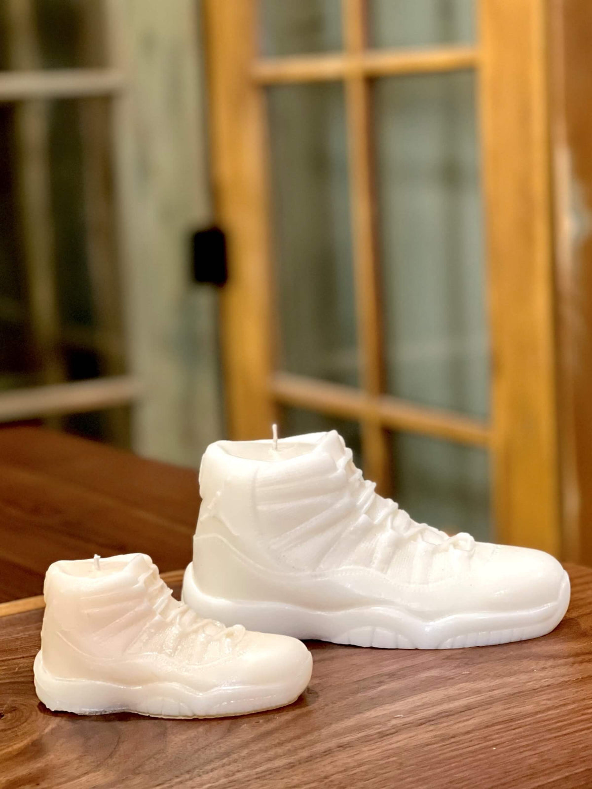 A large AJ-11 White Hightop Sneaker Candle sits on a wooden surface by glass-paneled doors. Hand poured to capture every detail, this candle adds a stylish accent to any space.