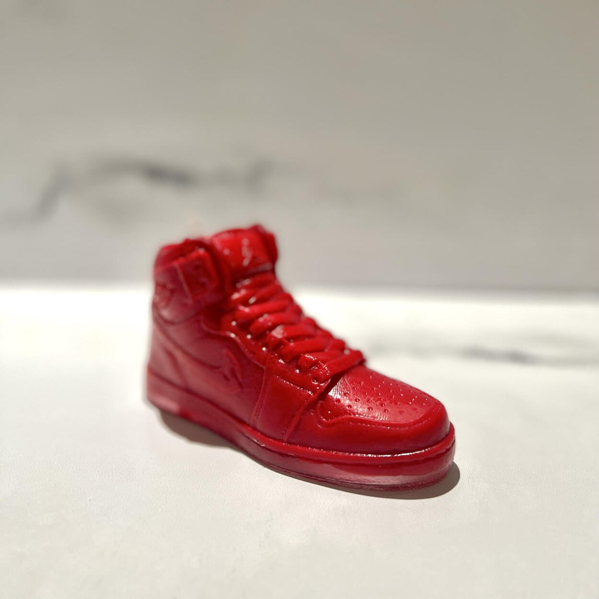 The AJ-1 Red Hightop Sneaker Candle – Large (currently on backorder) is a realistic, shiny red sneaker-shaped candle, hand poured and infused with Black Ice fragrance, displayed on a white surface with a blurred background.