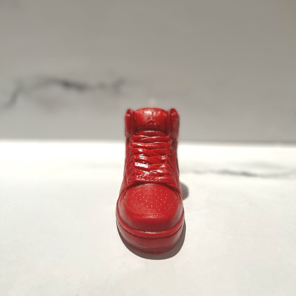 The AJ-1 Red Hightop Sneaker Candle – Small (On Backorder) is displayed on a white surface against a subtle gray marbled background.