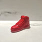 A hand-poured AJ-1 Red Hightop Sneaker Candle – Small (On Backorder), shaped like a bright red sneaker, rests on a white surface against a light gray and white background.