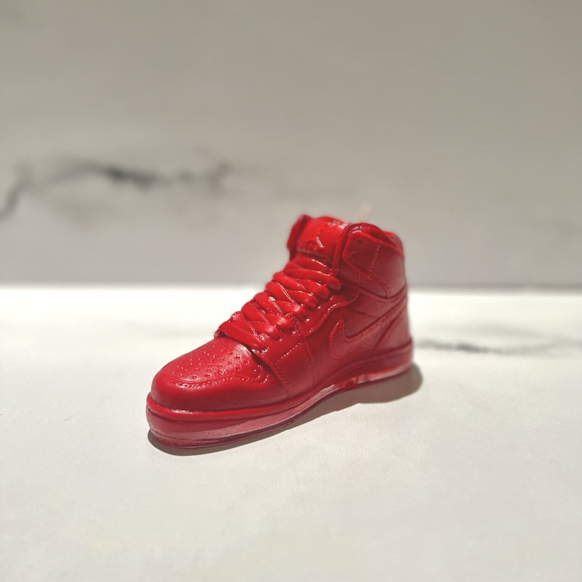 A hand-poured AJ-1 Red Hightop Sneaker Candle – Small (On Backorder), shaped like a bright red sneaker, rests on a white surface against a light gray and white background.