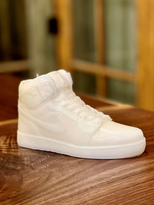 The AJ-1 White Hightop Sneaker Candle – Small (On Backorder), hand poured from all natural wax, rests on a wooden surface with a blurred backdrop of glass and wood panels.