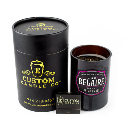 A Recycled Rare Rosé Champagne Candle by Custom Candle Co., crafted from a Luc Belaire bottle, sits beside a black container and branded matchbox on a white background.