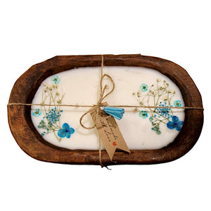 Oval white soap with blue pressed flowers and greenery in a rustic wooden dish, tied with twine and a tag—pairs perfectly with our Blue Floral Wood Candle – Floral Scented for a charming rustic centerpiece.