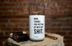 The "Thanks for Puttin’ Up with My Shit" candle, crafted from a recycled wine bottle, rests on a tree stump beside matches and a lighter, with a brick wall as the backdrop.