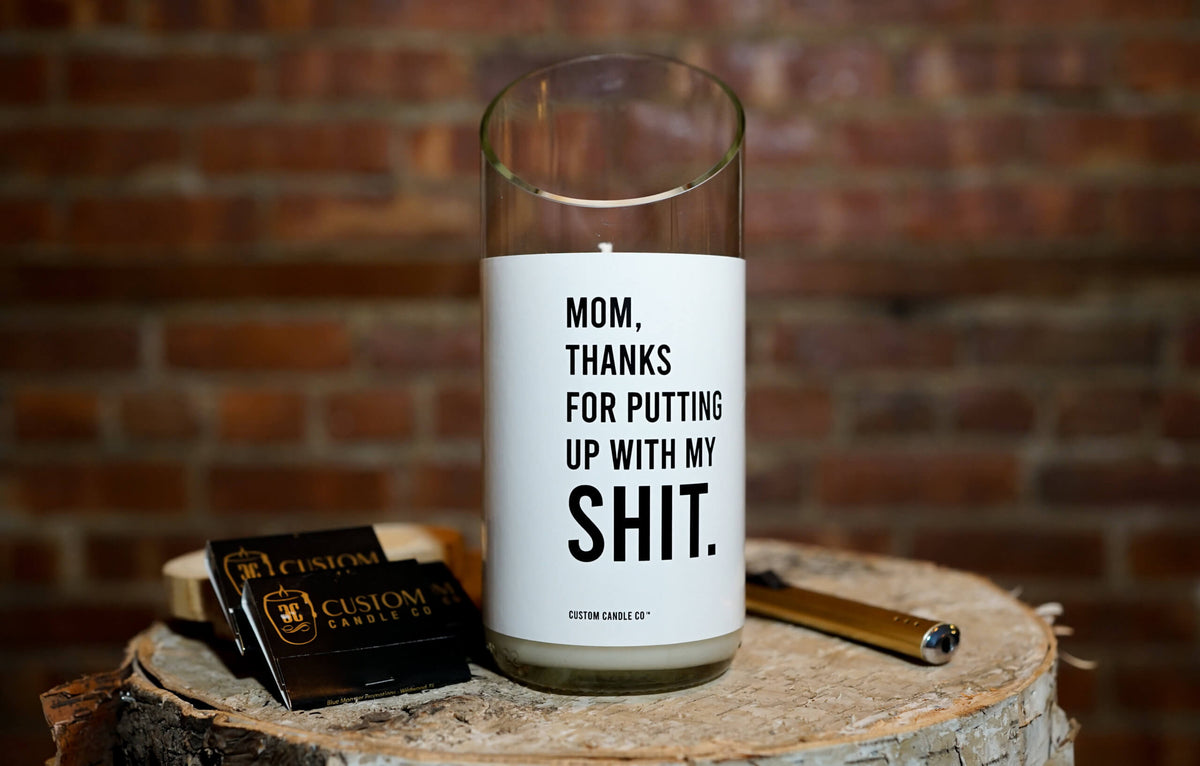 The "Thanks for Puttin’ Up with My Shit" candle, crafted from a recycled wine bottle, rests on a tree stump beside matches and a lighter, with a brick wall as the backdrop.