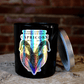 The Zodiac: Capricorn Candle—a black candle featuring holographic ram artwork and zodiac dates—rests on a wooden surface with its silver lid nearby, standing out elegantly against a brick wall backdrop.