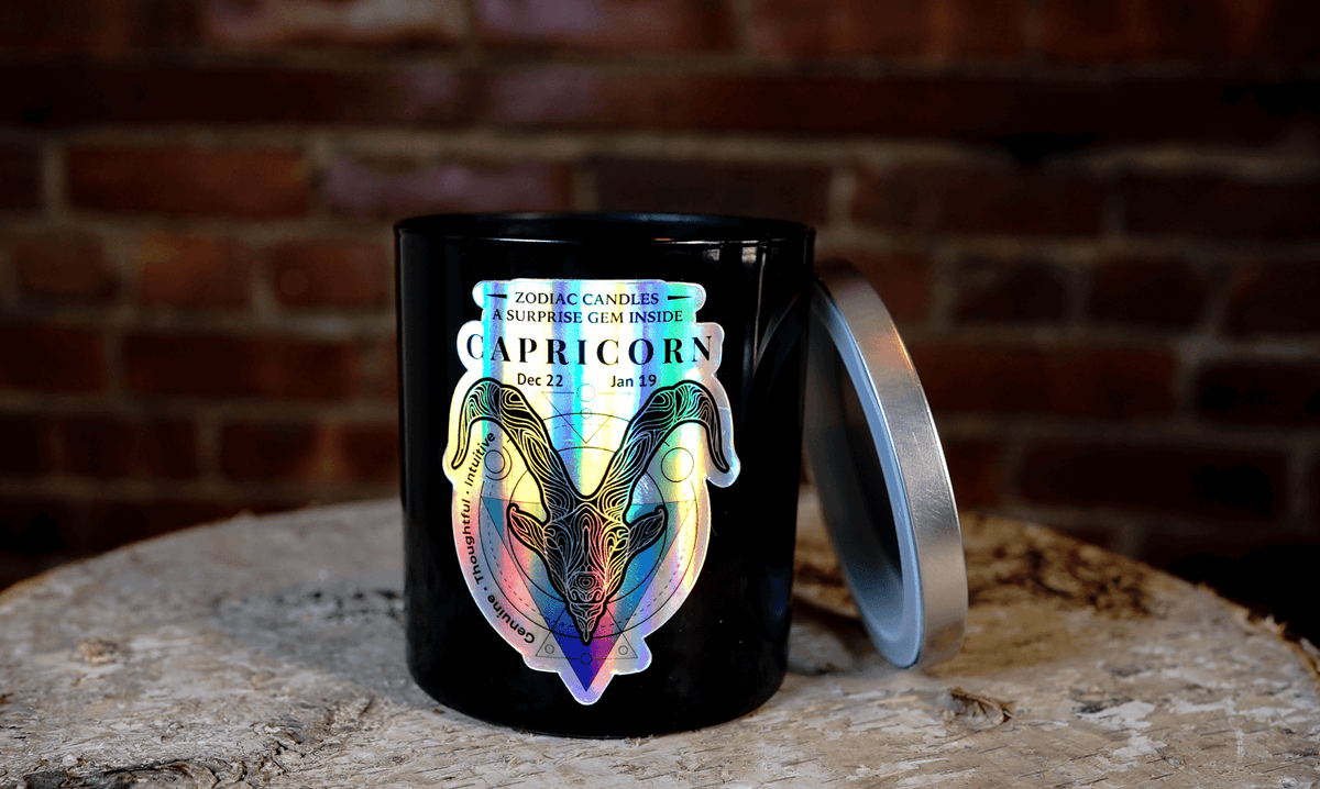 The Zodiac: Capricorn Candle—a black candle featuring holographic ram artwork and zodiac dates—rests on a wooden surface with its silver lid nearby, standing out elegantly against a brick wall backdrop.