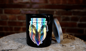 The Zodiac: Capricorn Candle—a black candle featuring holographic ram artwork and zodiac dates—rests on a wooden surface with its silver lid nearby, standing out elegantly against a brick wall backdrop.