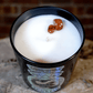 The Zodiac: Scorpio Candle—black glass jar with white soy wax and two small brown stones on top—sits on a rough stone surface, brick wall in the background.