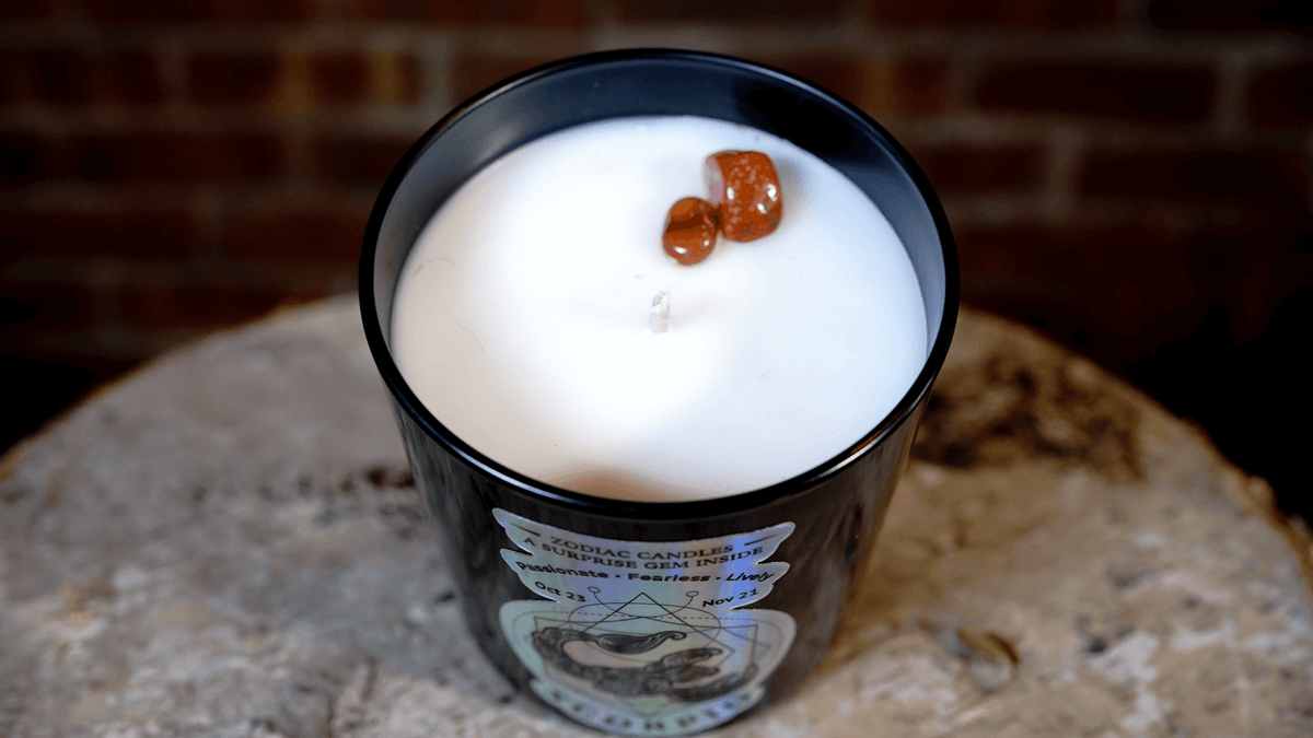 The Zodiac: Scorpio Candle—black glass jar with white soy wax and two small brown stones on top—sits on a rough stone surface, brick wall in the background.