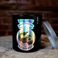 The Zodiac: Scorpio Candle features a black jar with a holographic design and partially removed silver lid, resting on wood against a brick wall—an ideal zodiac accent for any space.