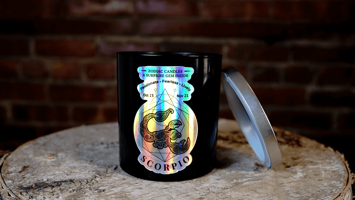 The Zodiac: Scorpio Candle features a black jar with a holographic design and partially removed silver lid, resting on wood against a brick wall—an ideal zodiac accent for any space.