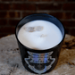The Zodiac: Taurus Candle is a black candle featuring the Taurus zodiac label and two stones set in white soy wax, placed on a wooden surface with a brick background.