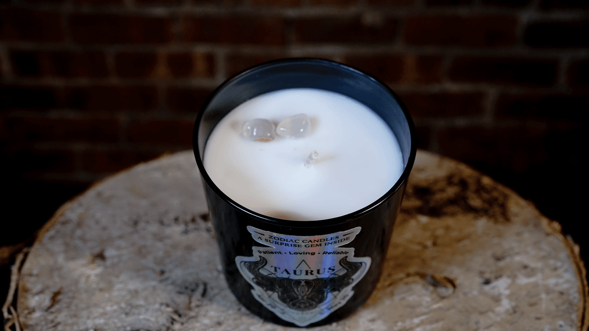 The Zodiac: Taurus Candle is a black candle featuring the Taurus zodiac label and two stones set in white soy wax, placed on a wooden surface with a brick background.
