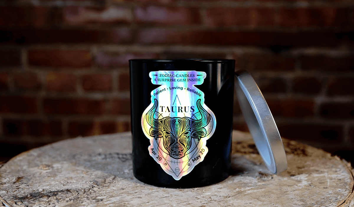 The Zodiac: Taurus Candle, featuring a holographic bull design and black jar, sits on a wooden surface with its silver lid beside it. A brick wall is visible in the background.