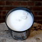 The Zodiac: Cancer Candle is a white soy candle in a black glass jar topped with two small stones, shown on a textured surface before a brick wall—ideal as a zodiac-themed gift.