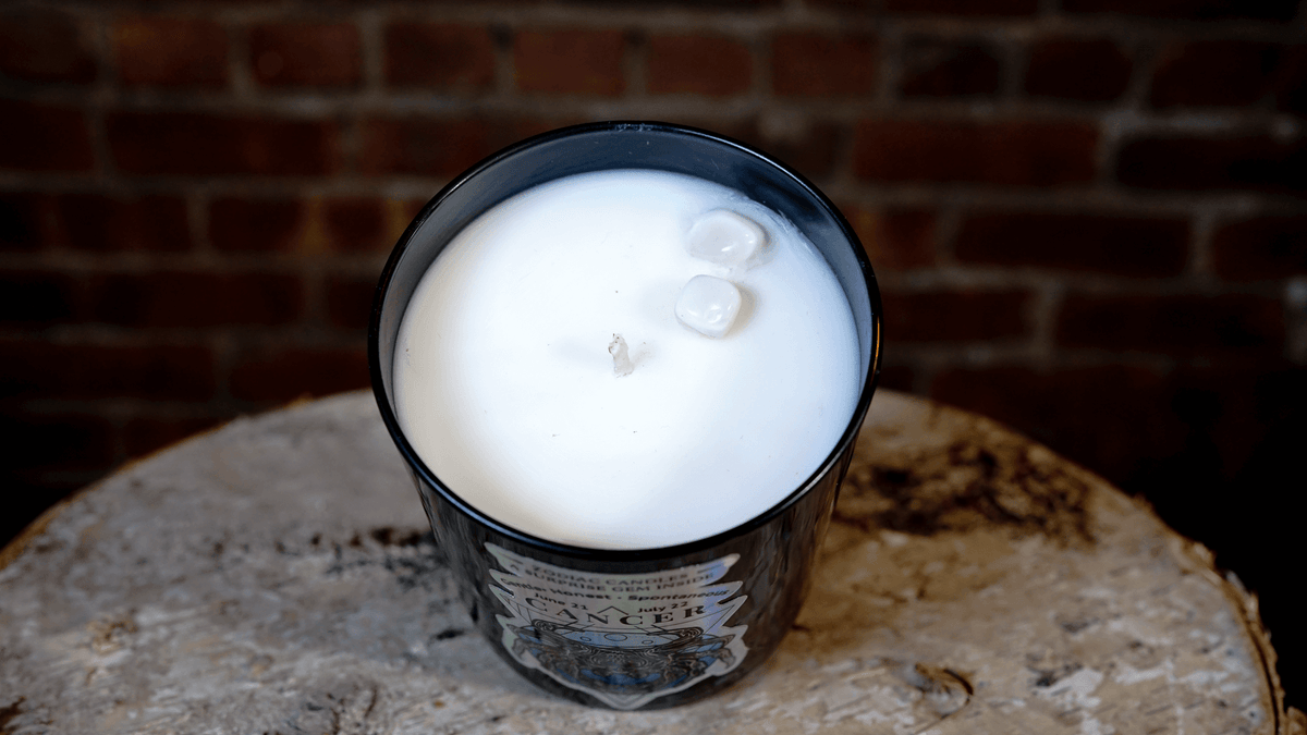 The Zodiac: Cancer Candle is a white soy candle in a black glass jar topped with two small stones, shown on a textured surface before a brick wall—ideal as a zodiac-themed gift.