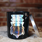 The Zodiac: Cancer Candle, a black jar featuring a crab illustration, zodiac dates, and personality traits, rests on a wooden surface with its silver lid propped nearby.