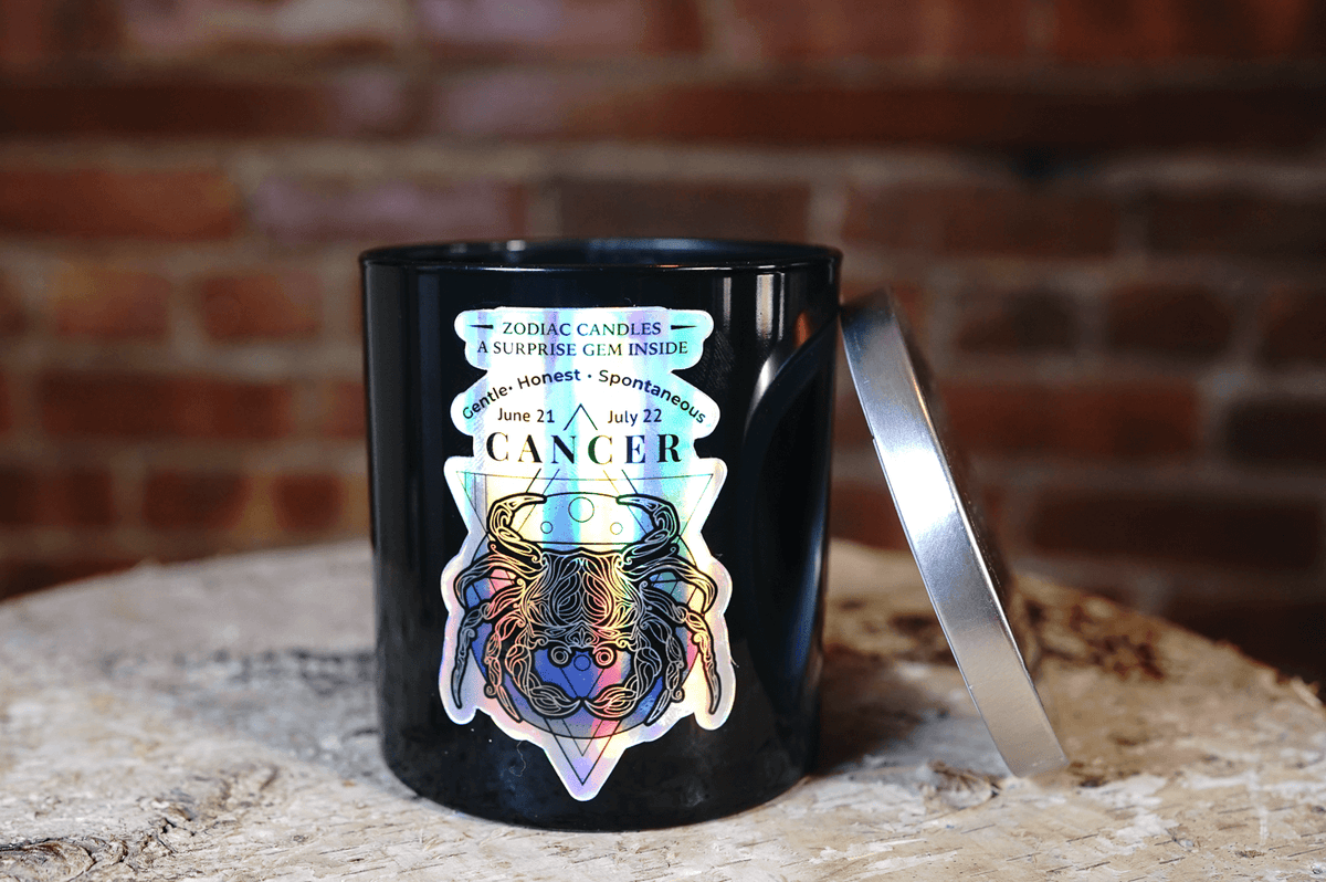The Zodiac: Cancer Candle, a black jar featuring a crab illustration, zodiac dates, and personality traits, rests on a wooden surface with its silver lid propped nearby.