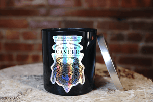 The Zodiac: Cancer Candle, a black jar featuring a crab illustration, zodiac dates, and personality traits, rests on a wooden surface with its silver lid propped nearby.