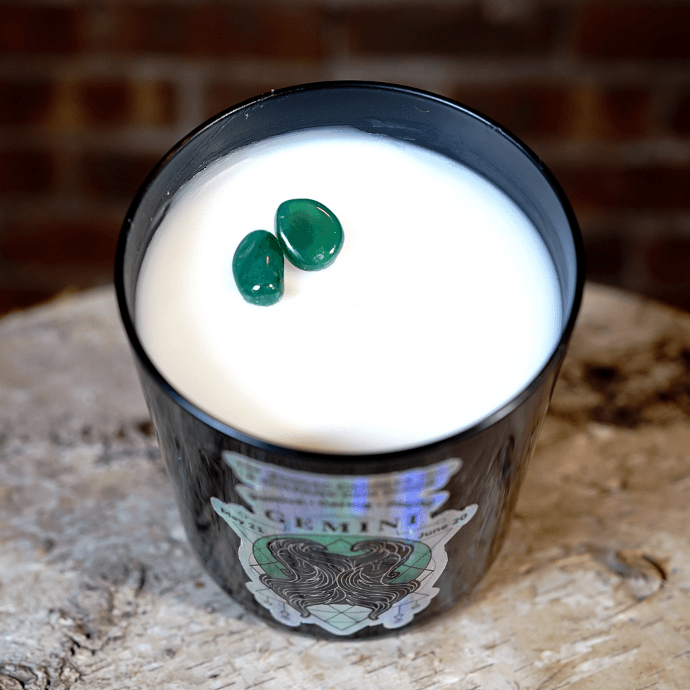 The Zodiac: Gemini Candle features a black jar filled with white natural soy wax and topped with two green stones, displayed on a wooden surface with a brick wall in the background.