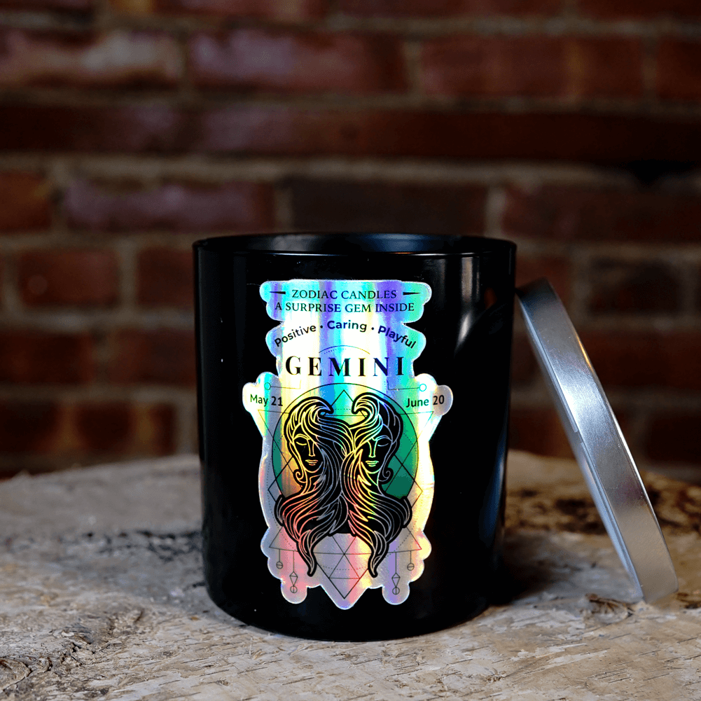The Zodiac: Gemini Candle, a black jar with a holographic design and astrological dates, rests on a rustic wooden surface beside its lid—a striking natural soy zodiac candle.