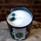 The Zodiac: Virgo Candle is a black natural soy zodiac candle with two green stones on top, displayed on a round, rough surface in front of a brick wall.