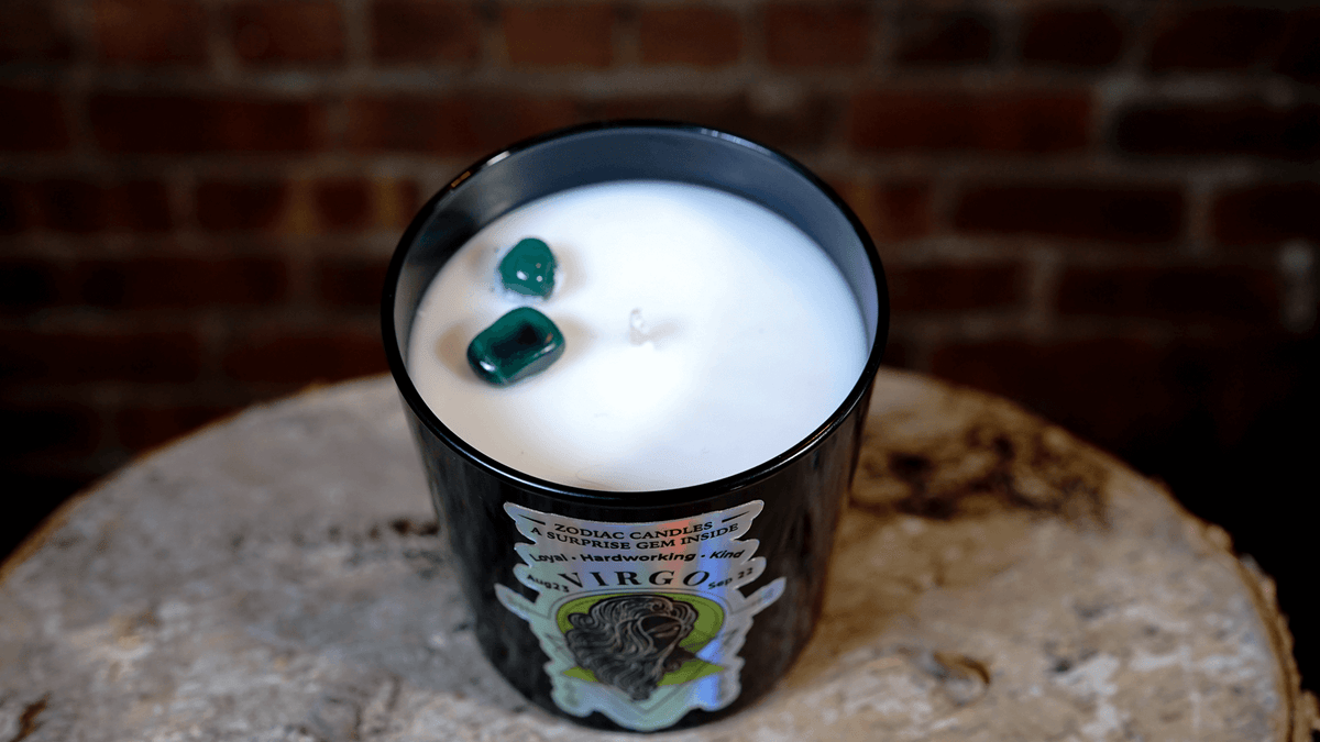 The Zodiac: Virgo Candle is a black natural soy zodiac candle with two green stones on top, displayed on a round, rough surface in front of a brick wall.