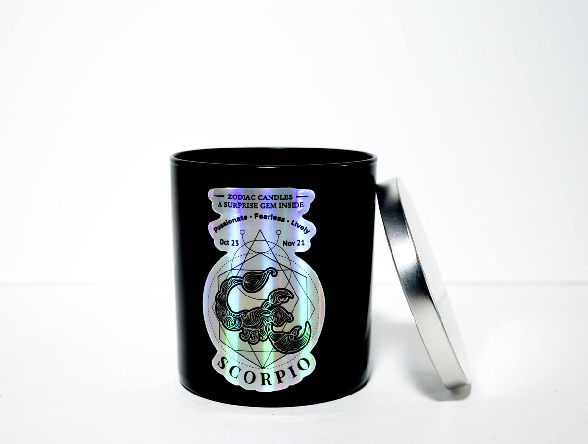 The Zodiac: Scorpio Candle features a black jar with astrological dates, a holographic design, and a silver lid, shown resting beside it on a white background.